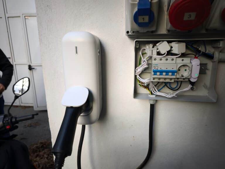 Trạm sạc xe điện (EV) gắn tường có cáp sạc màu đen được kết nối, bên cạnh bảng điện mở hiển thị hệ thống dây điện và cầu dao trên tường màu trắng.